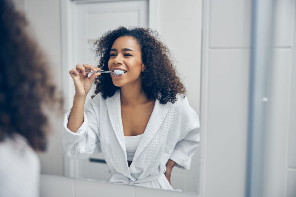 Woman in white robe brushing her teeth in bathroom mirror